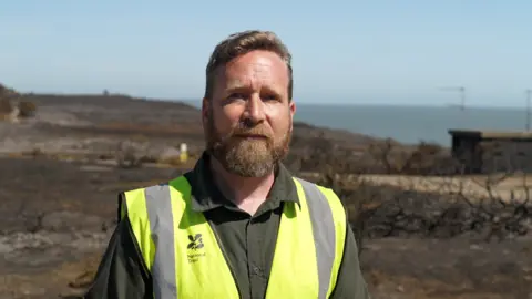 Jamie Niblock/BBC A bearded man wearing a high-vis vest with "National Trust" branding on it is standing in a field scorched black by fire. Blue skies and the sea can be seen on the horizon.