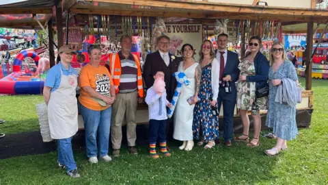 Andrew Stacey A wedding party lined up in front of a fairground ride, the central couple is the brode and groom. 