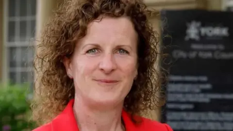 LDRS Claire Douglas, a woman with brown curly hair and a red jacket.