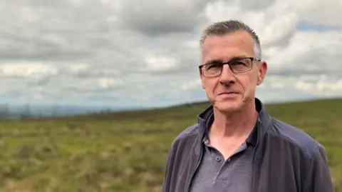 Mr McCabe has short grey and dark hair and is wearing rectangular glasss. He is smiling at the camera as he wears an unzipped fleece and a grey polo shirt. Behind him is the peatland which is mostly green.