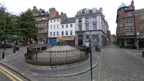 Google Streetview of a cobbled square with three and four storey buildings housing pubs and nightclubs. In the foreground is a round domed roof protruding from the square.