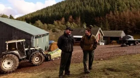 Laura and Mark Day, a couple in their 30s, are standing chatting in a farmyard dressed in dark trousers and jackets. He is wearing a cap and has a beard, she has short dark hair. 