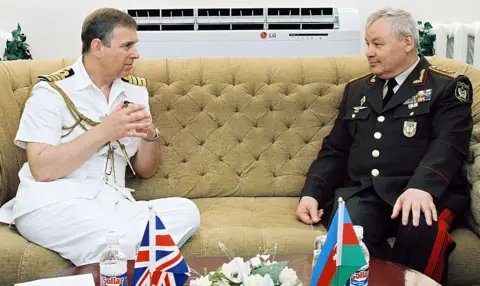 Getty Images Prince Andrew in Baku on a visit to Azerbaijan. Andrew wears a white naval uniform, he is talking to Colonel General Safar Abiyev, Azerbaijani defence minister, who wears a dark military uniform. They are sitting on a brown couch, with flags visible on the table in front of them. 