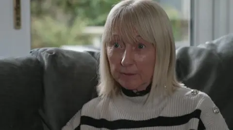 Frances Boyd, who has shoulder-length white hair and is wearing a white and black striped jumper, sits on a leather couch in front of a window. 