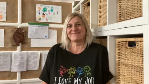 A woman with blonde hair is smiling and wearing a black T-shirt which reads 'I Love My Job'. She is standing against wicker storage baskets in a classroom. Behind her is a picture of the Gruffalo and children's pictures on the wall.