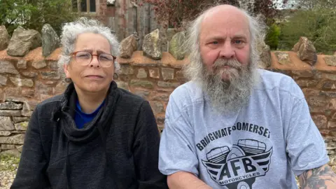 Two older people - a man and a woman - sit on a bench in front of a brick wall. The woman is in her 50s and has curly grey hair, and is wearing a black turtleneck and red-rimmed glasses. The man is in his 60s. He has a mostly bald head with wispy grey hair and a long beard. He is wearing light blue T-shirt with a motif on it and text that reads "Bridgwater, Somerset".
