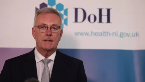 Health Minister Mike Nesbitt, with short grey hair, wearing glasses, a grey tie, white shirt and black suit. He is standing in front of a Department of Health poster. 