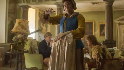 BBC/Bav Media A woman in a period drama costume, is looking at a distance with one hand raised. There are two men sitting in the background, speaking to each other.