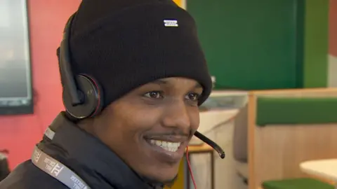 A young man wearing a black woolly hat and has a headset with microphone. He is smiling and is in a classroom setting.