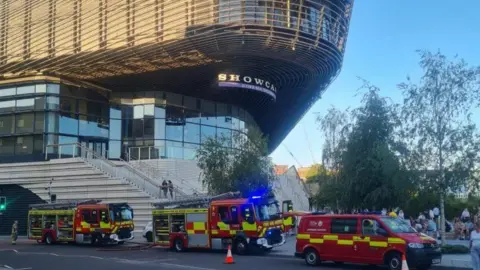 Fire engines and van parked up on a road outside a large shopping centre.