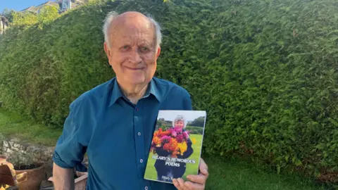 Mr Dennis Keam is standing in a garden holding the book. The front cover of the book has a picture of Bard Hilary Keam holding a bouquet of flowers. The title of the book reads Hilary's Humorous Poeams. Mr Keam is wearing a blue shirt and is standing in front of a green hedge.