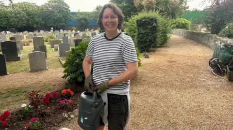 Lady in a cemetery wearing a striped t-shirt holding a large dark green watering can beside red and pink flowers.
