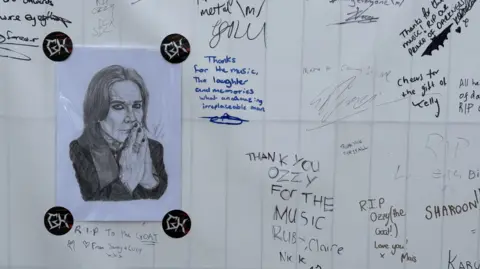 A close-up of a banner with written tributes to Ozzy Osbourne. A black and white pencil drawing of Osbourne with his hands pressed together near his mouth is also stuck to the board with black stickets.
