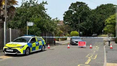 Police car on road with houses in the distance and a police cordon across.