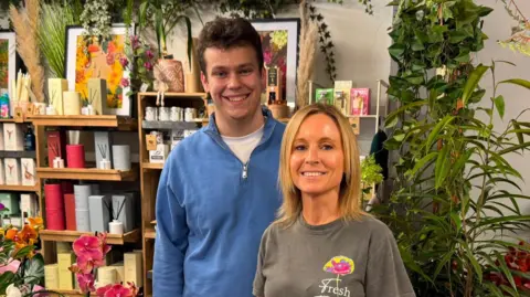 Jamie, who has short curly hair and a broad smile, wears a blue half zip fleece. His aunt Tracy, standing beside him, is a head shorter and has shoulder-length blonde hair. She is smiling too. Behind them are shelves filled with candles and pictures.