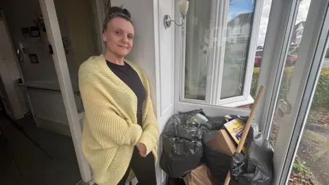 Tonia Dunn stands by her bags and bags of recycling in her porch area. She has her hair tied up and is wearing a black top and yellow cardigan. 