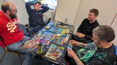 Rikh Hart Four men sitting around a table which is covered in cards