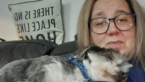 Karen Hawthornwaite A woman with grey/brown hair sits on a sofa smiling with a small fluffy dog asleep on her chest.
