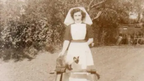 Black and white picture of Nora McCarthy standing in her garden wearing her nurses uniform. She is looking at the camera and her cat  is sitting in front of her.