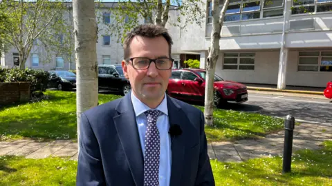 Devon and Cornwall Police Detective Inspector Dan Parkinson from the Devon and Cornwall Police Complex Fraud Team wears a blue shirt, blazer and tie and glasses and looks directly at the camera.