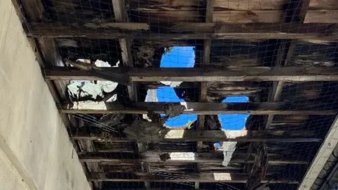Canopy above the Scott Cinema building with holes in it. There is a man walking along the pavement and there are buildings in the background.