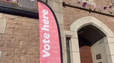 A red vertical sign, saying 'vote here' in white writing. It is displayed outside a granite building, with a wooden door visible on the right of the image.