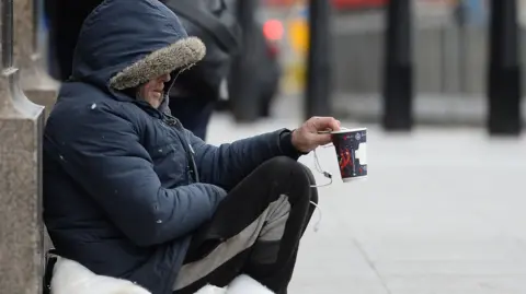 PA Media A man in a coat and tracksuit trousers holding up a coffee cup, asking for donations.
