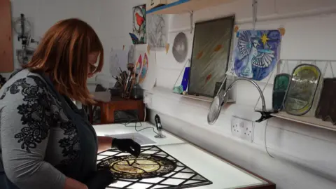 A woman with red shoulder-length hair leans over a light box. On the light box is a piece of stained glass. Colourful drawings are pinned to the wall in front of her.