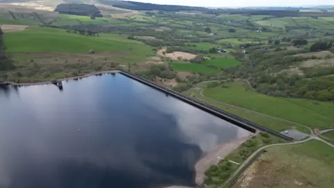Drone image of Crai Reservoir. A large body of water can be seen in the foreground, surrounded by hills, fields and trees