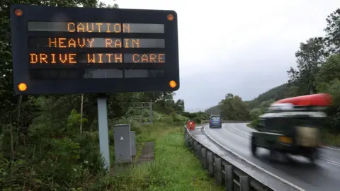 Reuters An electronic sign next to a road near Oban. It warns: "Caution. Heavy rain. Drive with care." A green LandRover with a red kayak on its roof is on the road next to the sign.