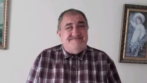 Paul Smith is smiling at the camera with a checked purple shirt buttoned up to his chin. He has light stubble and dark hair.