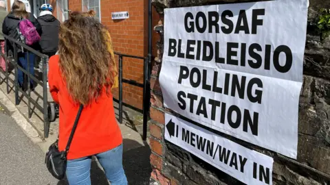 A woman with her back to the camera stood in front of a sign attached to a wall that says "Gorsaf Bledileisio, Polling Station"