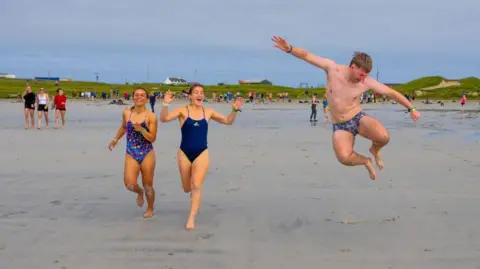Tiree Music Festival Participants run across a sandy beach after entering the water, with one person captured mid-jump while others gather in the background along the shoreline.