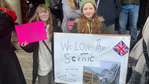 Cash Murphy/BBC Mia and Erin attending the protest with grandmother Susan. Susan is not in the image. Mia, on the left, is holding a small pink sign which says: "Save our Scenic" while Erin, on the right, is holding a bigger sign which says: "We love the Scenic". Erin's sign has an image of the rollercoaster and a taped on Union Jack flag.