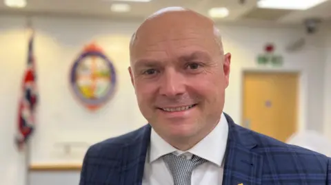 Andy Barnes has a shaved head and is wearing a navy suit jacket, white shirt and checkered tie. He is smiling inside the council chamber, where there is a union jack flag behind him.