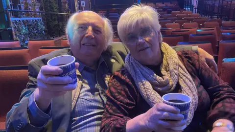 Shaun Whitmore/BBC Brian and Joyce Cooper sitting on seats in the theatre each holding a drink in a paper cup. Brian is dressed in a grey jacket and a blue pinstripe shirt. Joyce is wearing a red and black patterned outfit with a cream coloured scarf, and she has round glasses. They are both looking at the camera and smiling.