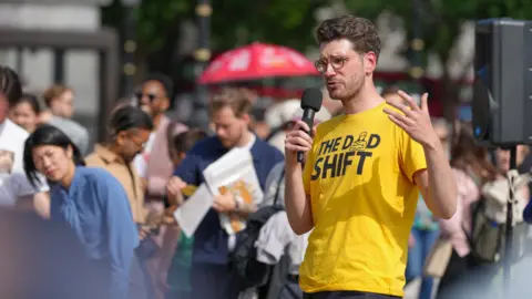 The Dad Shift Alex, wearing a yellow t-shirt saying 'The Dad Shift' standing in sunshine addressing a crowd with a microphone