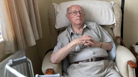 Family photo Garrod Evans sitting in a chair with his hands folded, with a walking frame and walking stick nearby