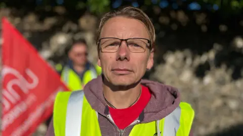 Martin Giles/BBC Andreas Kucher, a Unite the Union regional officer. He is standing outdoors on a picket line and a person is stood behind him waving a large red flag. Kucher is looking directly at the camera and is wearing a red T-shirt underneath a dark red zip-up hoodie. He is also wearing a yellow fluorescent hi-vis jacket.