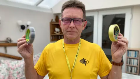 Keith Binks wearing glasses and a yellow t-shirt, holding up two types of sticky tape
