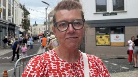 A person wearing glasses and a red and white top with a tie at the neck. They are stood nearby the road of the parade.