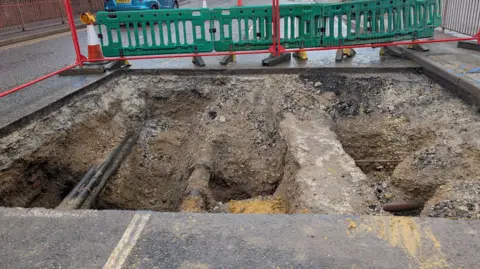 Southern Water Road with large hole dug out across surrounded by barriers - various pipes can be seen running across the length and width of the hole.