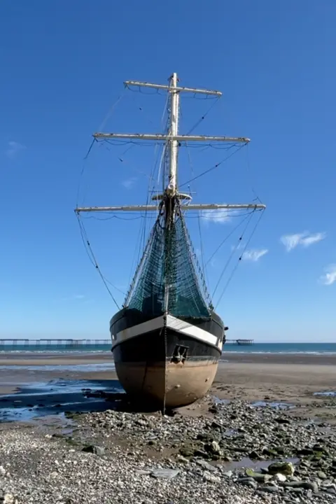 A front on view of the La Malouine grounded on a beach, with sand and rocks around it. It has a black hull with a white strip around it and tall masts with no sails. The tide is out and you can see a long iron pier in the distance, on a sunny day with blue skies and little cloud.