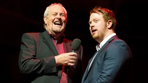 Two men standing side by side wearing shirts and blazers, one has short dark hair and one has short grey hair.