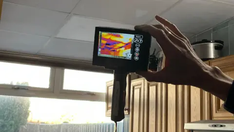 A close up of a hand holding an infrared camera to the ceiling of a kitchen, in front of a window. Yellow, purple and red can be seen on the screen.
