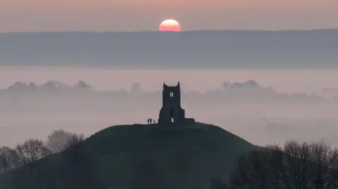 A silhouetted image taken from a distance, showing a group of people enjoying a misty sunrise over the monument at Burrow Mump in Somerset.