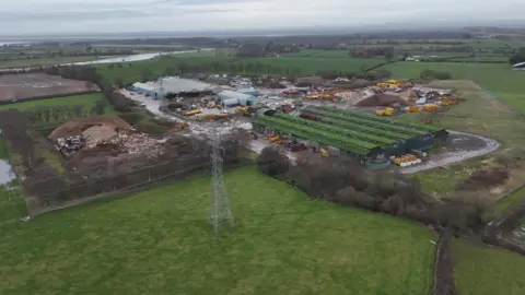 An aerial view of a large waste processing site. Sheds with green and grey roofs, piles of material and vehicles are visible at the site, which sits in the countryside, surrounded by fields with pylons.