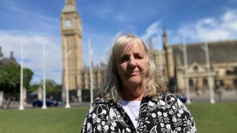 Joy Dove, vestida con un atuendo monocromático, está de pie en el césped frente al Parlamento, con el Big Ben de fondo.