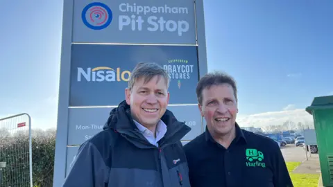 BBC David Hatherell is on the left, smiling and looking into the camera. He wears a grey waterproof jacket. Harling is on the right in a black polo top. Both are in front of the roadside for Chippenham Pit stop under a blue sky.