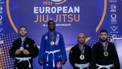 Gracie Barra Chelmsford Four men standing in front of a purple sign that reads "European Jiu-Jitsu IBJJF Championship". There are four men wearing medals. Three are wearing black robes and the other one is wearing a blue robe. The person wearing the blue robe is standing on a slightly elevated stand because he is the winner.